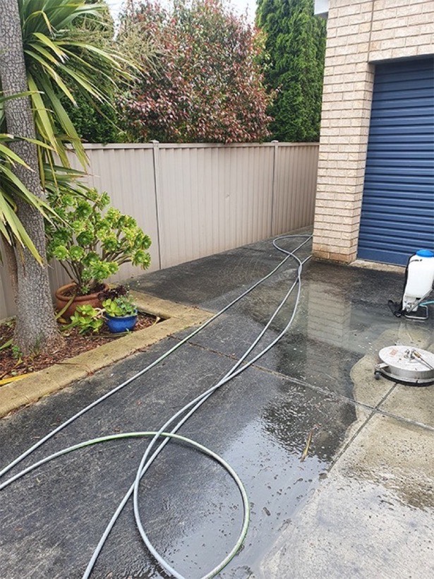A concrete path beside a colorbond fence part way through washing, wet and dark, with a surface cleaner and hose in frame